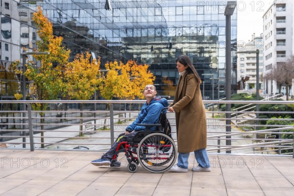 Woman pushing a smiling man in a wheelchair along an urban sidewalk during autumn, showcasing