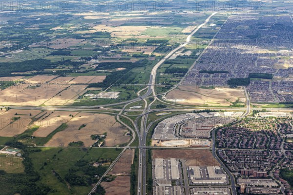 An aerial view shows a vast landscape of highways, fields and densely built suburbs under clouds,