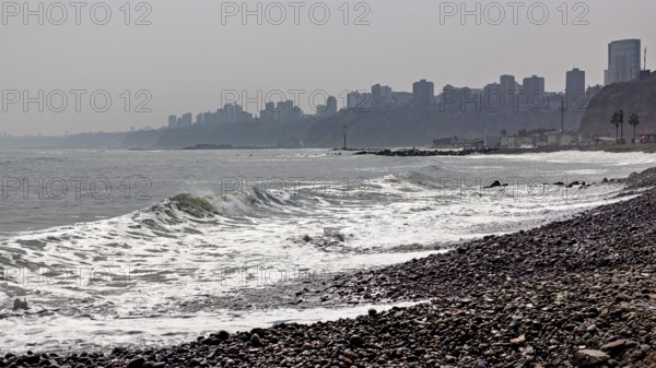 Pebble beach by the sea with waves, in the background skyscrapers of a city on a cloudy coastline,