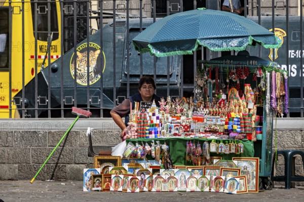 A woman sells colorful souvenirs at a street stand in a city with a bus in the background, a woman