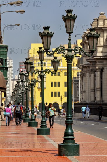 Row of green street lights along a busy walkway with architectural buildings in the background,