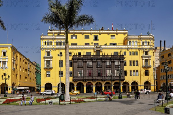 Wide square with yellow colonial buildings and palm trees surrounded by people and cars, The