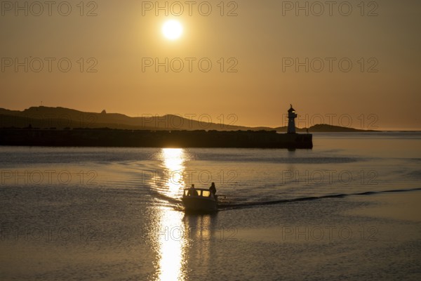 A small motorboat sails through the harbor entrance with lighthouse and midnight sun, Vardø,