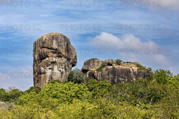 An impressive rock formation rises high above a green forest under a partly cloudy sky, the