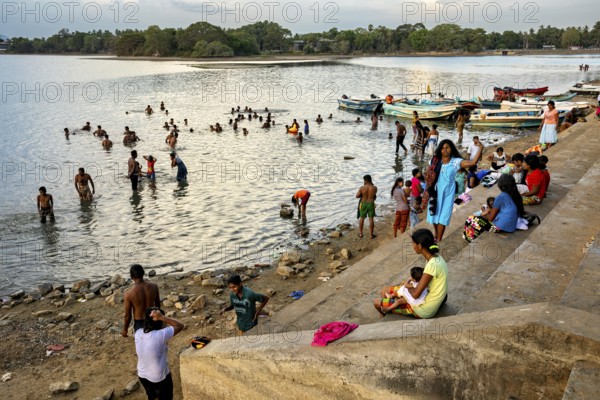 Children and adults on the riverbank, some bathe while others enjoy the view on the boats, people