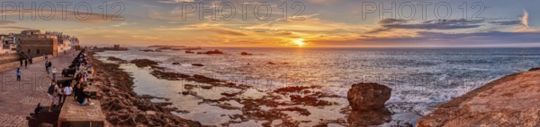 Panorama of a coastal promenade at sunset with rocks in the sea and a bright orange sky, Morocco,
