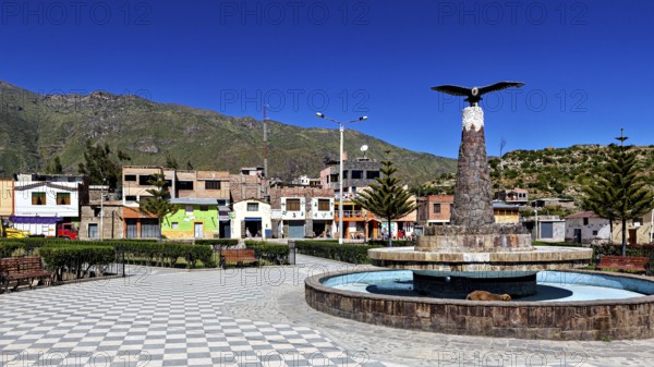 A public square with fountain and bird statue, surrounded by colorful houses and mountains, the