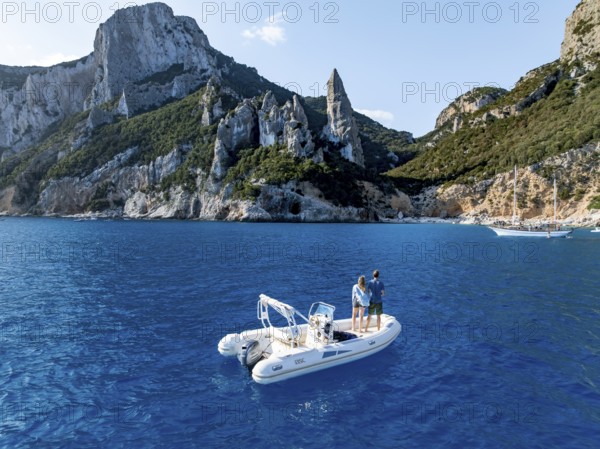 Couple on motorboat off picturesque rocky coast, cliffs with L'Aguglia pinnacle, blue sea and Cala