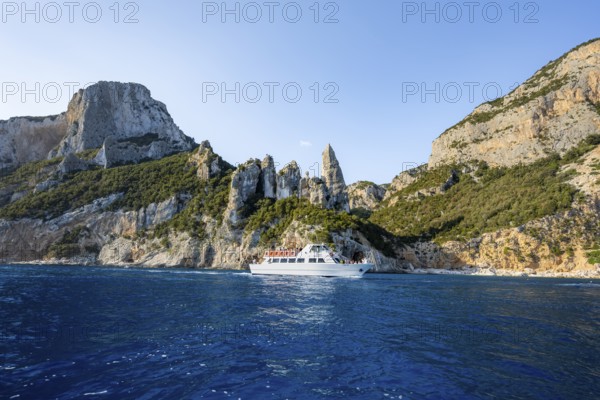 Excursion boat off picturesque rocky coast, cliffs with caves in morning light, L'Aguglia rock pin,