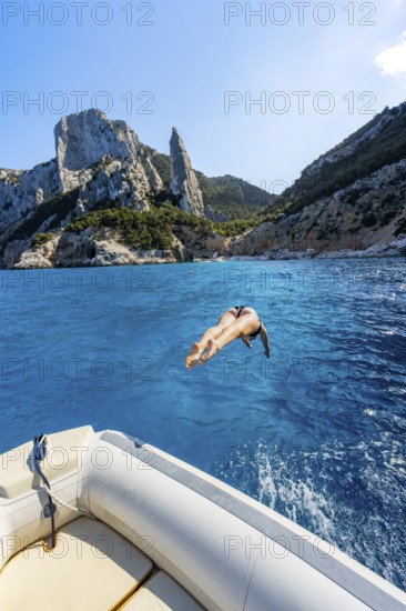Young woman jumping from a boat into the water, picturesque rocky coast, cliffs with L'Aguglia rock