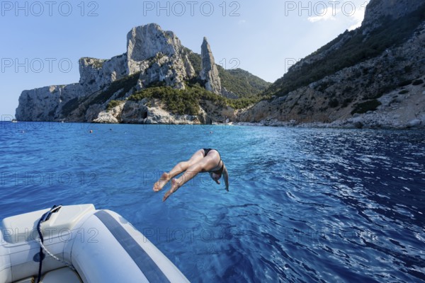 Young woman jumping from a boat into the water, picturesque rocky coast, cliffs with L'Aguglia rock
