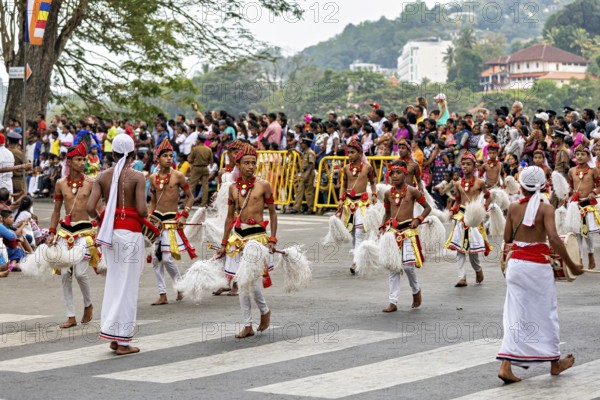 Children move across a street in traditional costumes, spectators watch, The Perahera Parade in