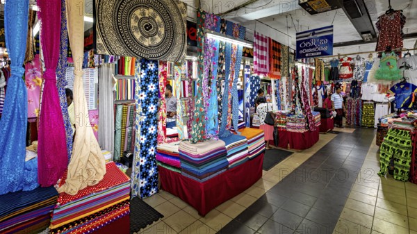 Various colorful fabrics and textiles are on display in a busy market hall, dealers in the market