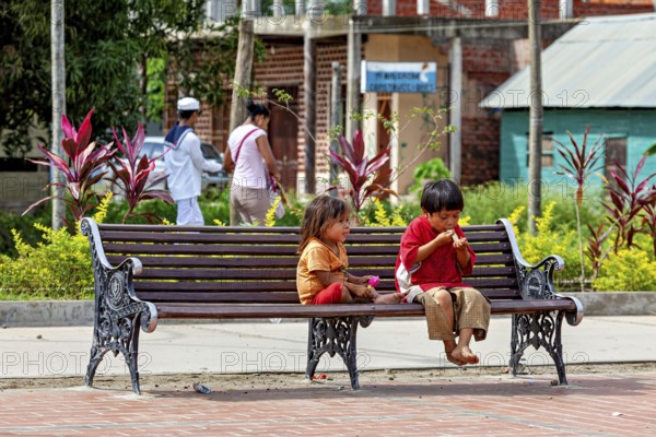 Two children sit on an outdoor bench while they eat. The surrounding area is green and peaceful,