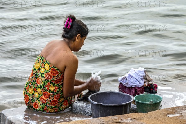 A woman is sitting by the river washing clothes with several buckets, a woman is washing at a lake