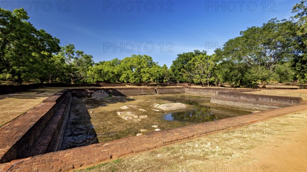 An empty brick water pool in a historic setting with green trees, the temples, dagobas, and pagodas