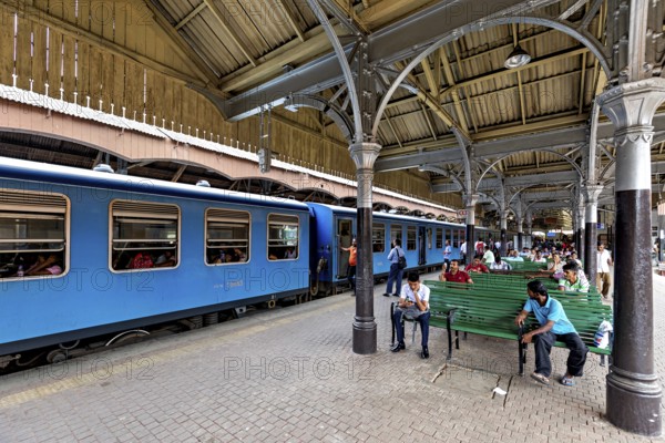 People sitting on green benches in the train station, blue train on the platform, covered