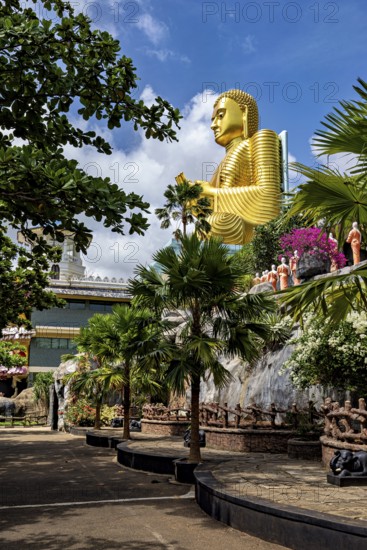 Large golden Buddha statue outdoors surrounded by trees and other sculptures under a blue sky, The