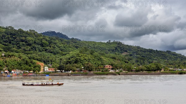A boat moves on a river in front of a wooded hill and under a cloudy sky, people on boats near