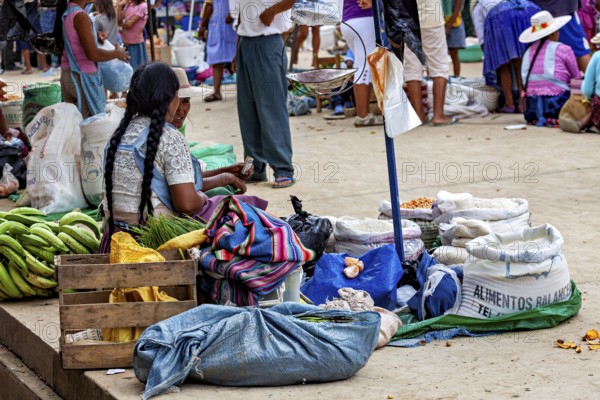 A woman sits in a market surrounded by sacks and bananas and observes the surrounding area, The