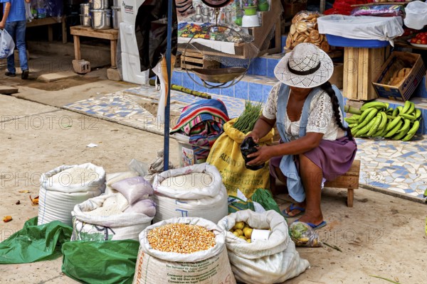 Woman wearing a hat sitting at the market and opening a bag of grain, The Rurrenabaque market in