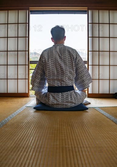 Young man wearing kimono sitting in traditional Japanese living room with tatami mats and shoji