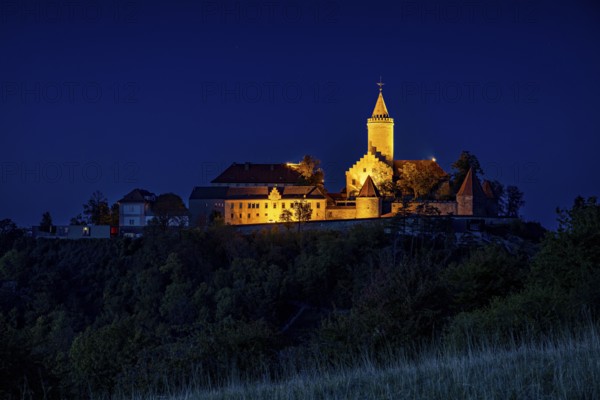 An illuminated castle is perched on a hill, surrounded by dark countryside and a deep blue night