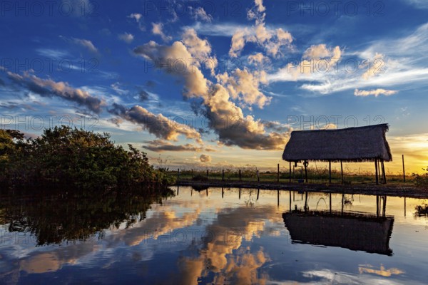 A thatched hut reflected in the water at sunset with dramatic cloud formations, The pampas swamps