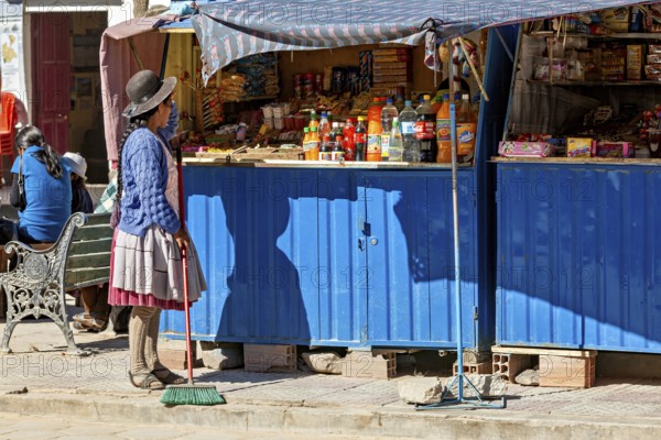 Woman in traditional dress sweeps in front of a blue market stall full of colourful bottles and