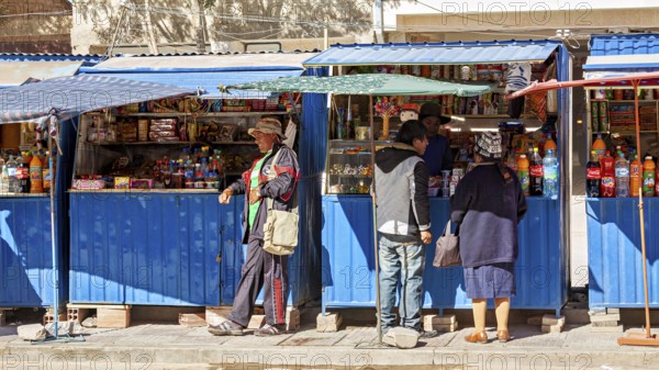 Street market with a blue stall, people buy colourful bottles and products under a blue sky, small