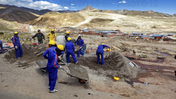 Workers in blue work clothes shovel earth on a sunny hilly landscape, surrounded by mountains and