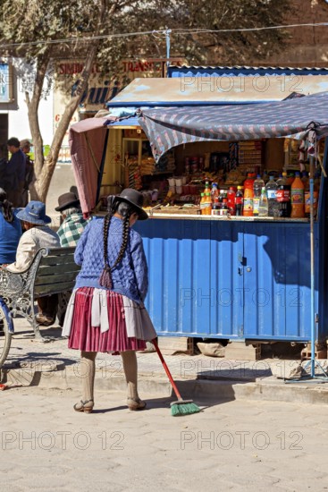 Woman in traditional dress sweeping in front of a blue stall on a busy street in the sunshine,