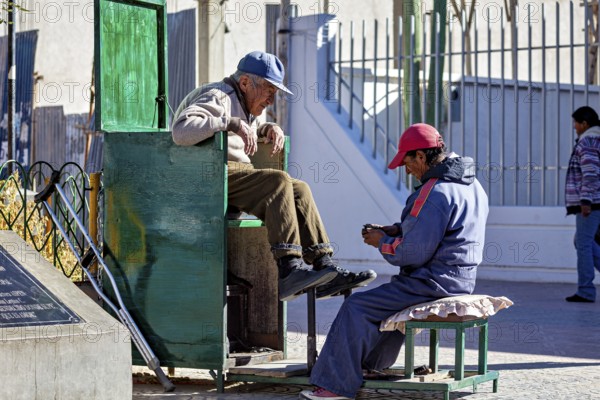 Shoe shiner concentrates on a man sitting on a green bench outdoors, show shiner in the city of