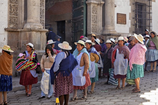Women in traditional dress stand on a busy cobbled street in front of a historic building,
