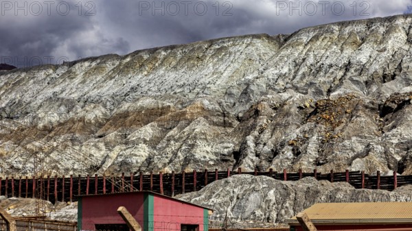 Industrial landscape with buildings at the foot of steep rock faces under a cloudy sky, slag heap