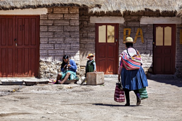 A woman in traditional dress carries bags while children and another person sit in the background,