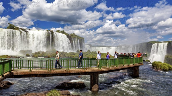 People walk on a bridge in front of an impressive waterfall under a blue sky, The Iguazu Falls