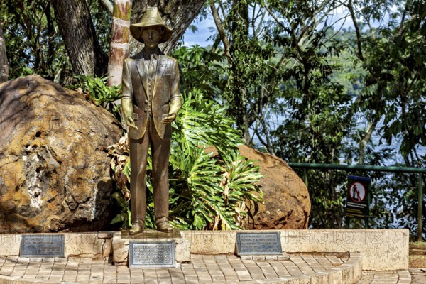 Bronze statue of a man wearing a hat and suit, set in a natural environment with trees and plaques,