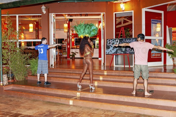 Woman in feather headdress walks past a group of spectators, samba dancers to entertain tourists at