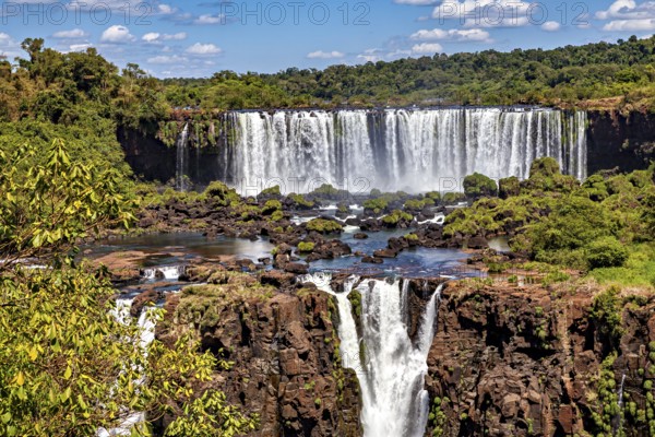 Steep waterfalls cascade over rocks, surrounded by dense greenery and a cloudy sky, The Iguazu