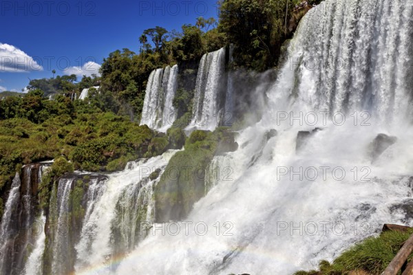 Massive waterfalls create dense spray and a rainbow, surrounded by green forest and blue sky, The