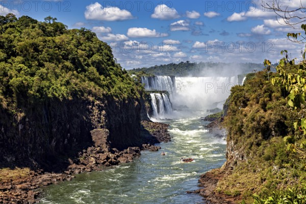 Majestic waterfalls flow between lush vegetation and steep rock faces under a clear sky with white