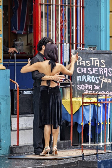 A couple dances tango in front of a colourful building, hugging each other, next to a menu board,