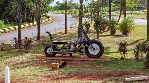 A metal motorbike bust stands in a park with green spaces and trees, placed on a pedestal, A