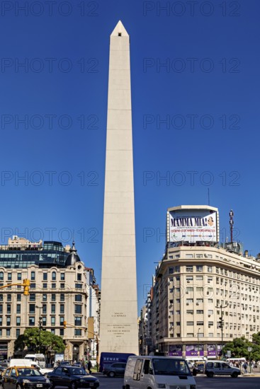 A tall obelisk stands between modern buildings under a clear blue sky, The Great Obelisk in the