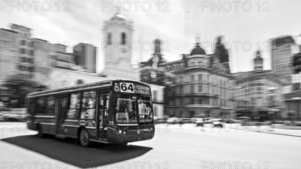 A bus drives through a busy city with historic buildings in the background in black and white, Bus