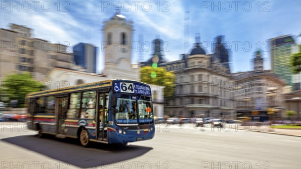 A colourful bus drives through a busy city with historic buildings and a blue sky in the