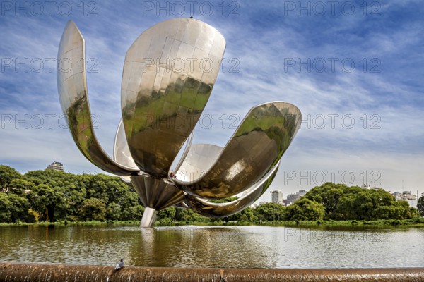 Large metallic flower with reflective surface in a park with water and green foliage, The lotus