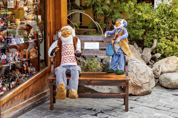 Two dolls sit on a bench in front of a shop window, surrounded by plants and wooden surfaces,