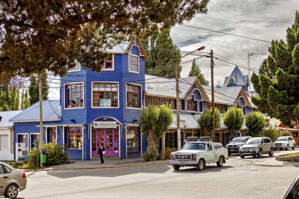 A blue, two-storey building in a busy street with parked cars and trees, street scene in El Chalten